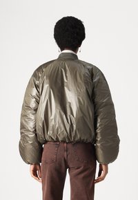 Olivgrüne Pufferjacke mit glänzender Außenseite, geripptem Kragen und elastischem Saum, getragen über rostfarbenen Jeans. Glatte Textur und voluminöse Form.