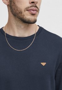 Temno moderna sweatshirt z teksturirano tkanino; ima majhen trikotni usnjen pečat in zlato verižico.