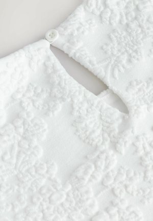 Prenda blanca con un patrón floral texturizado. Presenta un escote en forma de llave y un cierre de un solo botón, con un detalle de tela en capas.