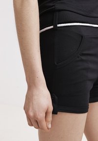 Pantalones cortos de tela negra con un diseño ajustado, bolsillos laterales y una cintura con cordón blanco, con un acento de botón en el dobladillo.