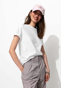 T-shirt blanc à manches courtes avec des poignets roulés, associé à un pantalon taille haute rayé léger gris et violet. La casquette est rose clair avec un motif brodé.
