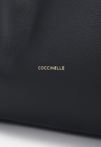 Surface en cuir texturé noir avec le nom de la marque embossé en or "COCCINELLE" centré dessus.