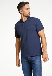 Mörkblå pikétröja med klassisk krage, korta ärmar och en liten logotyp på bröstet; kombinerad med denimjeans.