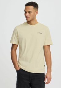 Camiseta de algodón de un beige claro con mangas cortas y cuello redondo. Presenta un pequeño logo negro en el lado izquierdo del pecho. Textura suave.