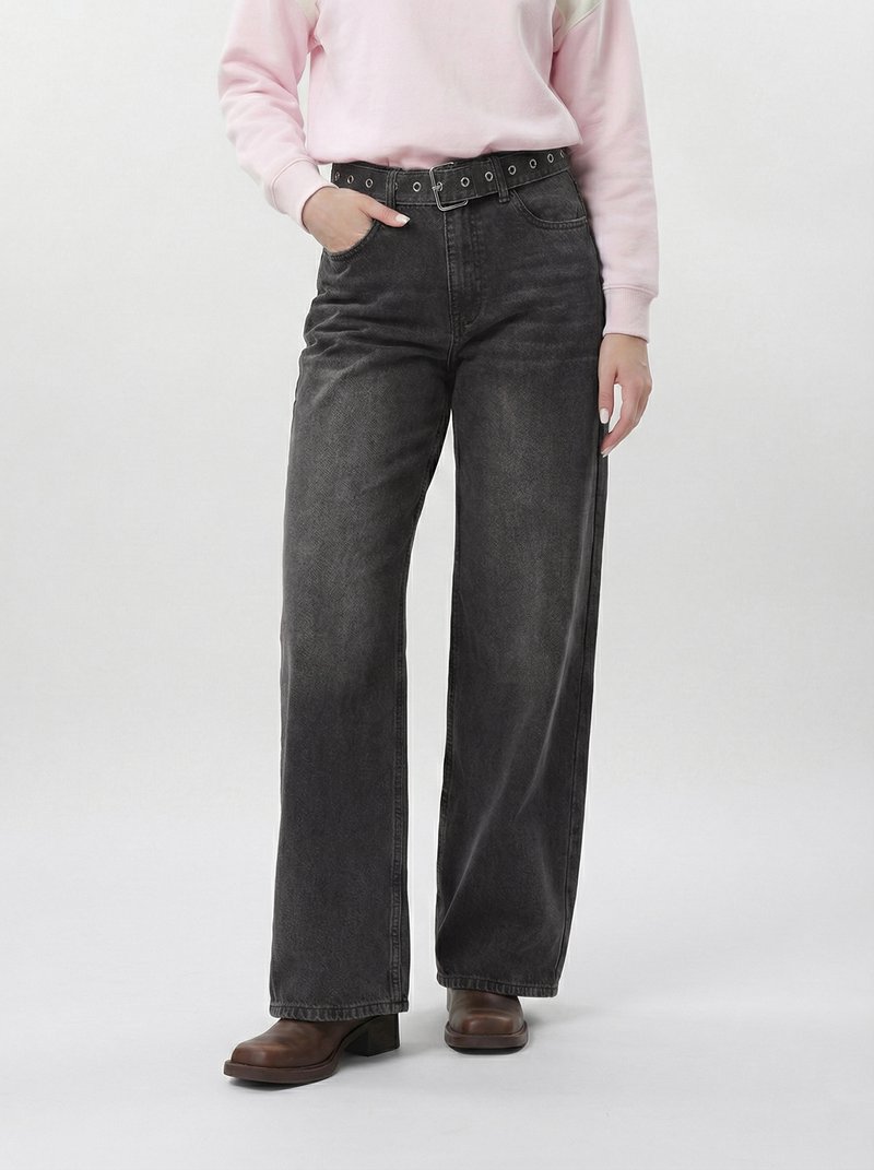 Egy személy, aki kopott fekete, magas derekú, széles szárú farmernadrágot visel, fekete övvel, rózsaszín pulóverrel és barna, sarokkal ellátott csizmával, egyik kezét a zsebében tartva áll.