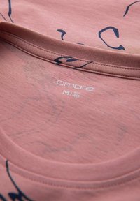 T-shirt in cotone rosa con scollo rotondo, decorata con motivi astratti neri e un'etichetta in tessuto che mostra "Ombre" e i dettagli della taglia.