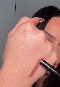 Une main trace une ligne brun rosé sur le dos de l'autre main avec un crayon de maquillage noir.