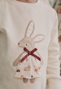 Cremefarvet sweatshirt med broderet kanindesign med quiltede ører, iført en kjole med blomsterprint og en mørkerød sløjfe.