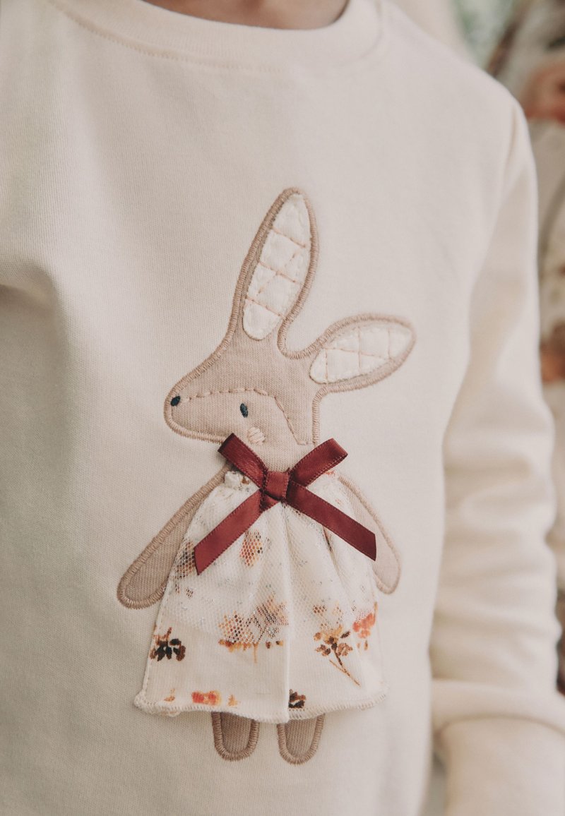 Sahnefarbener Pullover mit einem gestickten Hasendesign, das gesteppte Ohren, ein Kleid mit Blumenmuster und eine kastanienbraune Schleife trägt.
