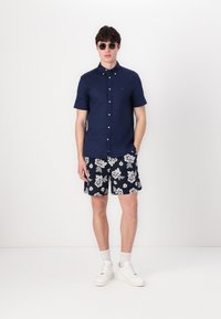Marineblå kortermet skjorte med knapper, marineblå shorts med blomstermotiv, hvite joggesko og solbriller. Avslappet sommerantrekk med kontrasterende detaljer.