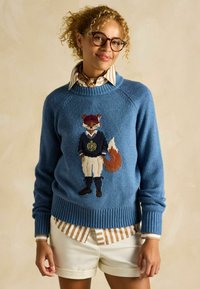 Pulover tricotat albastru cu un design brodat de vulpe îmbrăcată în echipament sportiv, cu manșete și tiv cu nervuri. Purtați peste o cămașă cu dungi.