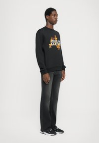 Μαύρη μπλούζα με το κείμενο "VERSACE JEANS COUTURE" σε λευκό και ζωηρό πορτοκαλί γραφικά, συνδυασμένη με σκούρα γκρι τζιν και μαύρα sneakers.