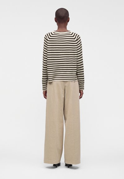Personne debout, dos à la caméra, portant un haut à manches longues rayé noir et blanc et un pantalon large beige sur un fond uni.