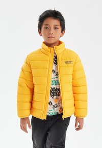 Gul puffjacka med hög krage och dragkedjestängning, med ett quiltat mönster. Bärs över en t-shirt med grafiskt tryck och färgglada accenter.