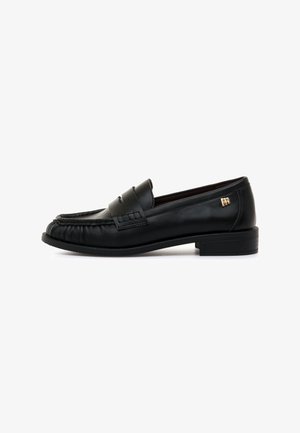 Mocassin noir en cuir avec bride pour pièce, couture surélevée sur la pointe, petit talon et petit logo doré sur le côté près du talon.