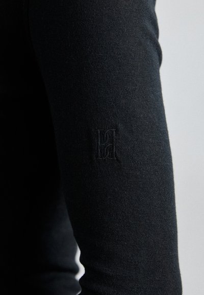 Must musta pikkade varrukatega rõivas, millel on sile tekstuur ja millel on varrukal väike tikitud logo. Kangas tundub veniv ja keha lähedalt istuv.