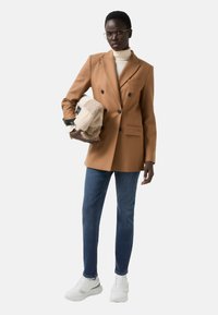 Hnědý dvouřadý blejzr s černými knoflíky, zkombinovaný s tmavými džínami a bílými teniskami. Drží béžovou a krémovou texturovanou tašku.