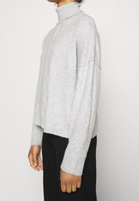 Maglione grigio a collo alto con una texture a coste, vestibilità relax, spalle scese e un orlo leggermente accorciato. Indossato sopra pantaloni neri.