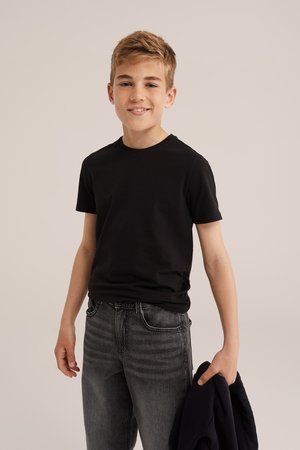 Băiat zâmbind, purtând un tricou negru și blugi gri, ținând o geacă neagră în mâna dreaptă, pe un fundal simplu.