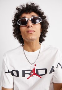 Ochelari de soare cu rame argintii și lentile întunecate, purtați cu un tricou alb cu un logo roșu Jordan și un colier cu lanț argintiu.
