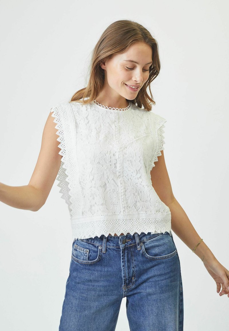 Top bianco in pizzo con bordi ondulati, scollatura rotonda e motivi floreali, abbinato a jeans di denim blu.