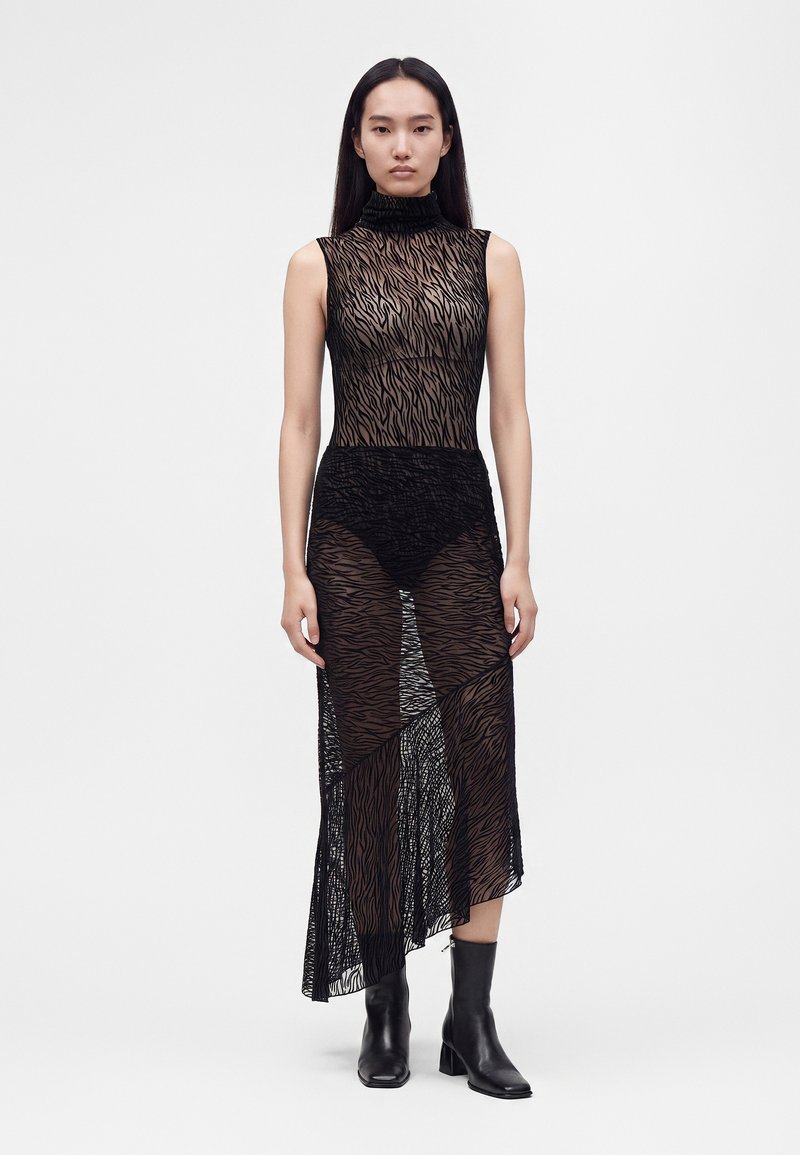 Femme aux longs cheveux foncés portant une robe noire transparente sans manches à imprimé animalier avec un col montant et des bottines noires sur fond blanc.