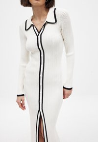 Raitareunukjole i hvit med svart krage og trim. Har frontknapper og spalte på siden. Lange ermer og tettsittende silhuett.