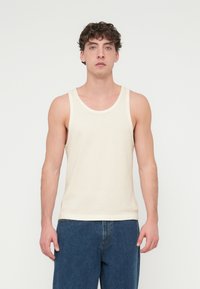 Μπεζ βαμβακερό τοπ χωρίς μανίκια με υφή, που διαθέτει φαρδιά στρογγυλή λαιμόκοψη και λεπτές τιράντες στους ώμους. Φοριέται με μπλε τζιν.