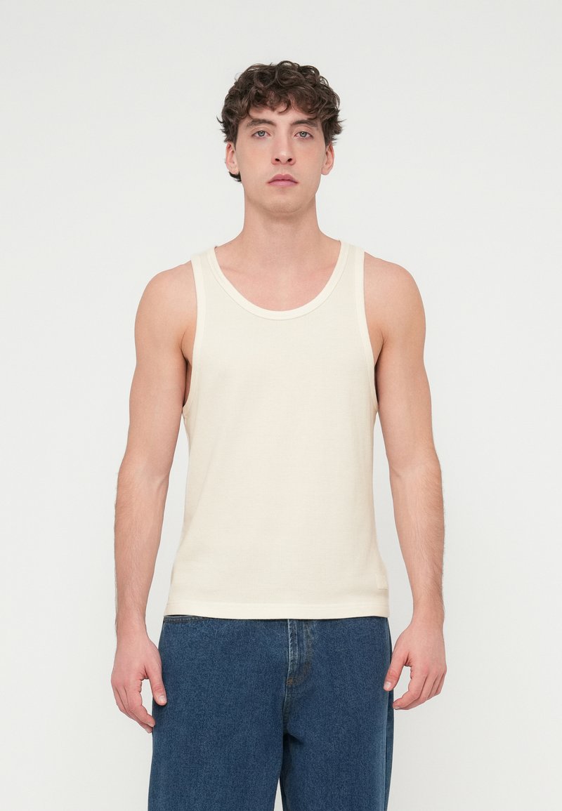 Μπεζ βαμβακερό τοπ χωρίς μανίκια με υφή, που διαθέτει φαρδιά στρογγυλή λαιμόκοψη και λεπτές τιράντες στους ώμους. Φοριέται με μπλε τζιν.