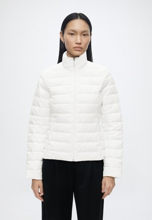 Vrouw met lang zwart haar draagt een witte doorgestikte jas, volledig dichtgeritst, en zwarte broek, staand tegen een effen witte achtergrond.