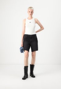 Débardeur blanc ajusté avec un logo noir, associé à un short en denim noir. Le modèle porte des bottes noires jusqu'aux genoux et tient un morceau de denim. Fond uni.