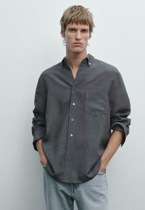 Young man with light brown hair wearing a dark gray button-up shirt with rolled sleeves and light blue jeans, standing against a plain wall.