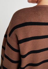 Pull marron avec des rayures horizontales noires, fabriqué en matériau doux, présentant un large col et une coupe décontractée, mettant en valeur une texture lisse.