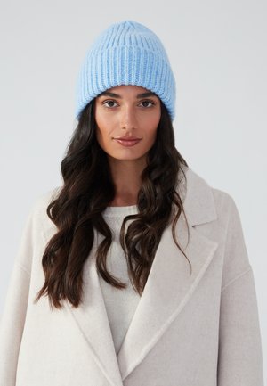 Strickmütze in Hellblau mit vertikaler Rippstruktur, getragen mit einem beigen Mantel und einem hellen Pullover, was einen lässigen Lagenlook präsentiert.