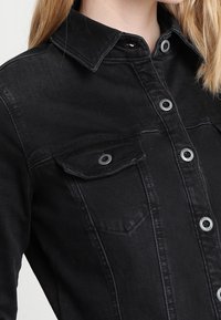 Svart denimjacka med krage, två bröstfickor och silverfärgade tryckknappar. Har en texturerad, något blekt yta.