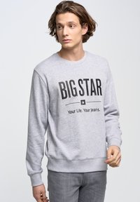 Siv sweatshirt z okroglim ovratnikom z izrazitim črnim napisom "VELIKA ZVEZDA" in sloganom. Mehak material, rebrasti manšeti in hem, oblečen z sivimi jeans hlačami.