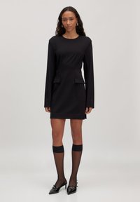 Vrouw die staat in een aansluitende zwarte jurk met lange mouwen en klepzakken, transparante knielange sokken en zwarte puntige hakken met gespen.