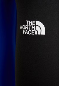 Czarne legginsy z elastycznego materiału o gładkiej strukturze. Posiadają wyraźne, białe logo The North Face na dolnej części nogi.