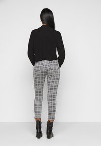 Frau mit langen Haaren steht, trägt einen schwarzen, kurzen Pullover, schwarz-weiß karierte Hosen und schwarze Stiefeletten vor einem weißen Hintergrund.