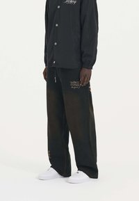 Chaqueta negra con botones a presión, llevada sobre pantalones de pierna ancha de color verde oscuro y marrón con un patrón que presenta bordados naranjas, y zapatillas blancas.