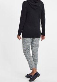 Zwarte gebreide trui met een capuchon, gecombineerd met grijs-witte geruite broek. Schoenen zijn glanzend marineblauw met accenten.