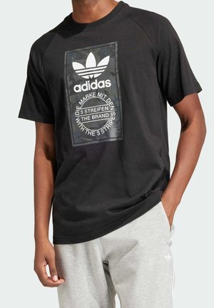 Mann trägt schwarzes Adidas-T-Shirt mit Logo und Schriftzug, kombiniert mit hellgrauen Adidas-Sweatpants, steht mit einer Hand in der Tasche.