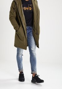 Oliwkowa parka z kapturem, czarny t-shirt z grafiką, zniszczone jasnoniebieskie dżinsy i czarne sneakersy z fakturowanymi detalami.