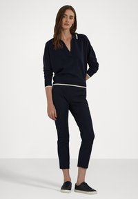 Navy gebreide pullover met een V-hals en crèmekleurige afwerking, gecombineerd met strakke navy broek en zwarte sneakers, tegen een eenvoudige witte achtergrond.
