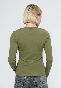 Haut à manches longues vert olive avec une texture côtelée et une encolure légèrement arrondie au dos. Présente une petite étiquette logo au niveau du cou.