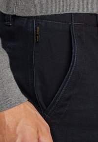 Donkerblauwe denim broek met een gladde textuur, voorzien van zijzakken en een klein logo-label. Een grijze gebreide trui is gedeeltelijk zichtbaar.