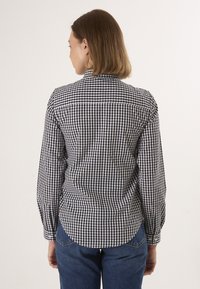 Camicia a quadretti bianco e nero con maniche lunghe, colletto e orlo curvo. Il tessuto appare leggero con una texture liscia.