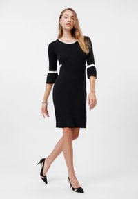 Black dress with three-quarter sleeves, white trim at neckline and sleeves, fitted shape, ribbed texture, and knee-length hemline.