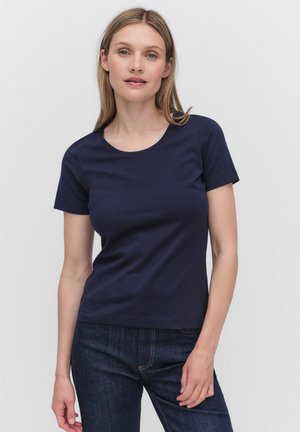 Vrouw met steil blond haar, gekleed in een effen marineblauw T-shirt met korte mouwen en donkerblauwe jeans, staande tegen een witte achtergrond.