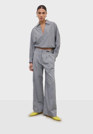 Kobieta w dużej szarej koszuli w prążki, szerokich szarych spodniach i jasnożółtych szpiczastych butach stoi na jednolitym tle.